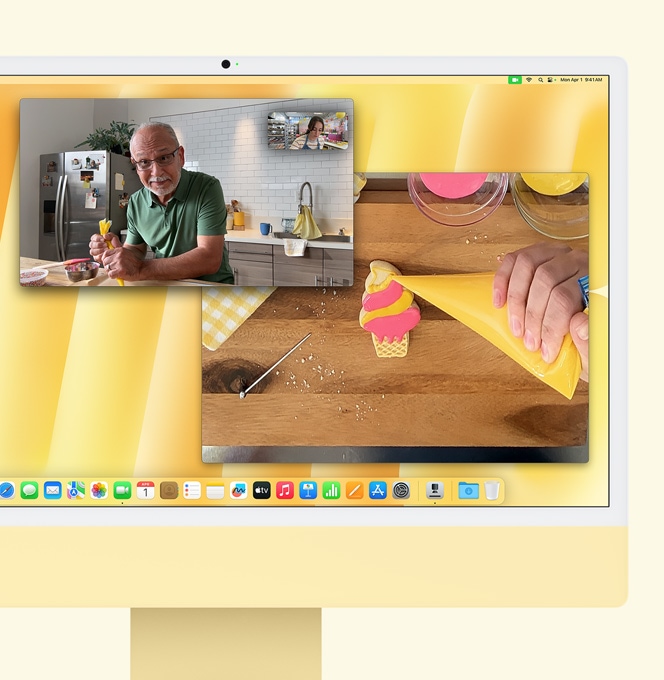 Κίτρινος iMac. Demo για το Desk View. Η οθόνη δείχνει τον αρτοποιό σε βιντεοκλήση με ένα άλλο άτομο. Στη μία οθόνη εμφανίζεται ο φούρναρης και το πρόσωπο του άνδρα. Στην άλλη οθόνη εμφανίζονται τα χέρια του αρτοποιού που γαρνίρουν ένα μπισκότο
