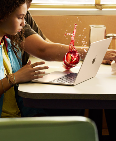 Eine Person greift in einem Restaurant nach etwas und verschüttet ein gekühltes Getränk über einem geöffneten MacBook