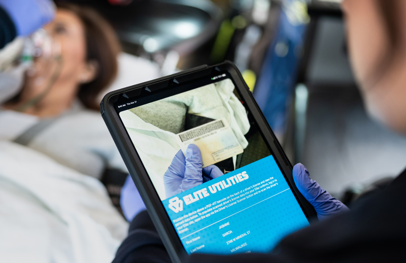 De Elite Utilities-app is geopend op iPad en een paramedicus scant het rijbewijs van een patiënt, terwijl de patiënt op een brancard ligt