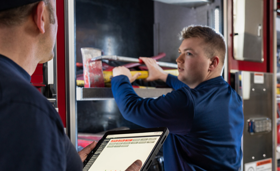 Twee medewerkers inspecteren gereedschap en apparatuur van een brandweerwagen, de een houdt een iPad vast terwijl de ander inspecteert 
