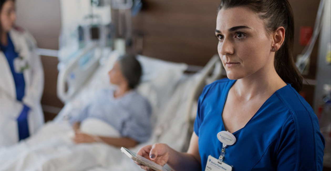 Une infirmière saisit du texte sur son iPhone tandis qu’une autre soignante discute avec une personne hospitalisée.