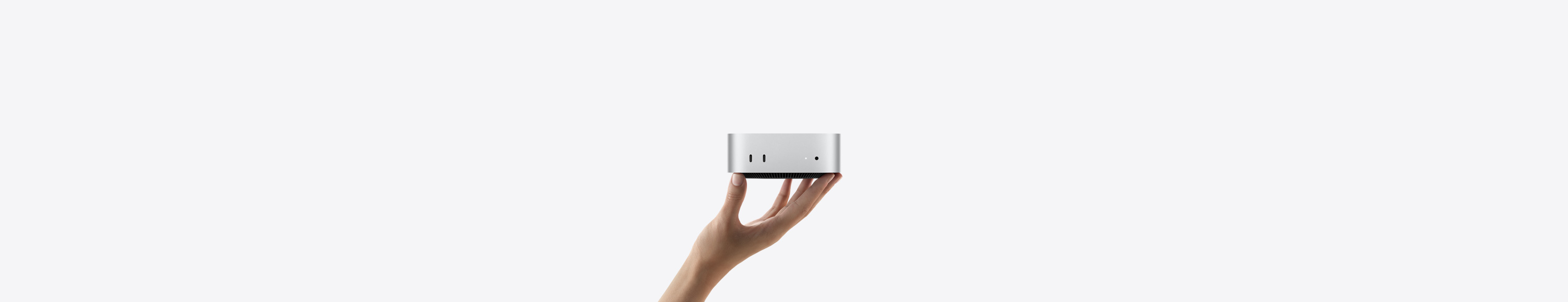 لقطة أمامية لجهاز Mac mini مسنود على رؤوس أصابع يد ممدودة للأعلى، الجهة الأمامية تعرض اثنين من منافذ ثندربولت، ضوء مؤشر التشغيل ومقبس لسماعات الرأس، قاعدة سوداء مدببة في الأسفل، سطح مسطح، جوانب مدورة، حواف مستقيمة، لون فضي