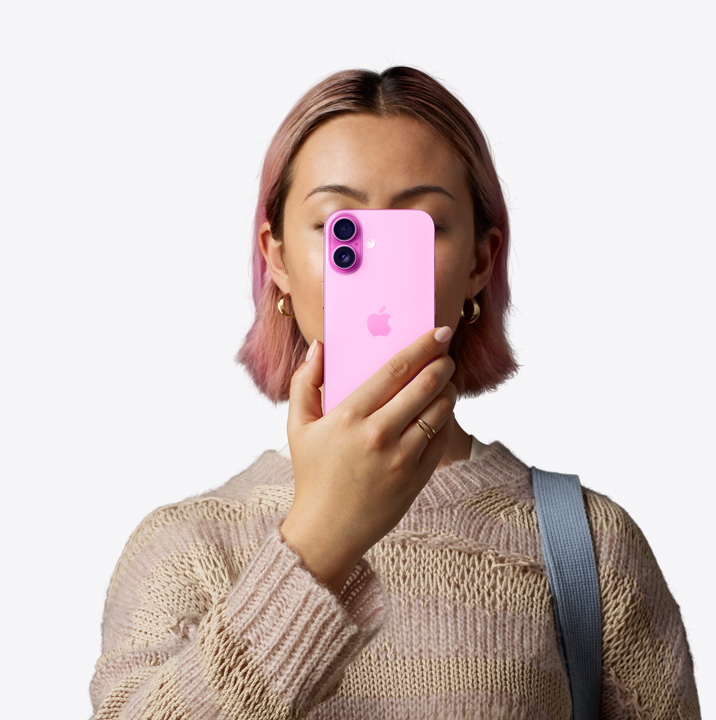 Žmogus laiko rožinės spalvos „iPhone16“ priešais veidą, matoma įrenginio galinė dalis, dviejų kamerų sistema kairiajame viršutiniame kampe.